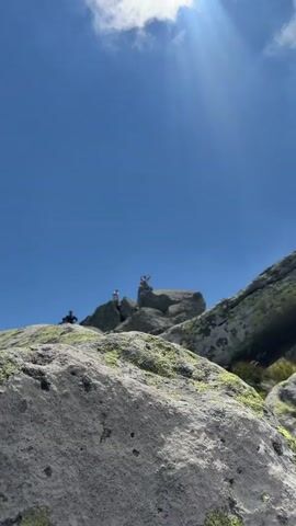 The video opens with a low-angle shot of a clear blue sky with a sun flare in the upper right corner. In the foreground, large, rough, grey rocks with patches of green moss are visible. In the mid-ground, several people are standing on top of a rocky outcrop. One person, in the center, is wearing a white tank top and grey shorts, and is flexing their arms. Another person is standing to their left, and a third person is partially visible to the left of the center person. The video then zooms in on the person in the white tank top and grey shorts who continues to flex their arms, then lowers them and turns their head to the side. The camera angle remains low, emphasizing the height of the rocky perch against the vast expanse of the sky.