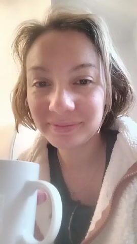 A woman is centered in the frame, holding a white mug with black paw prints. She has fair skin, hazel eyes, and light brown hair with blonde highlights. She's wearing a dark gray top and a white, fluffy zip-up hoodie. The woman takes a sip from the mug. The background is a blurred white wall.