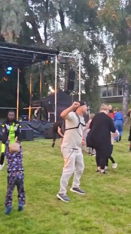 A group of people are dancing on a grassy area outside a building. There is a stage with speakers and lighting equipment on the left side of the frame. Several individuals are actively dancing, with some appearing to be following choreographed moves. The building in the background has several windows and a light-colored facade. The overall atmosphere appears to be a casual outdoor gathering or event.