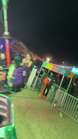 The video shows a blurry shot of a carnival ride at night. The ride appears to be spinning, with bright lights in the background. There are people riding the ride, and one person is seen holding a phone and taking a selfie. The ground is covered in grass and there are some barriers around the ride.