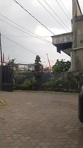 THE VIDEO SHOWS A VIEW OF A GATE AND THE AREA IMMEDIATELY BEHIND IT. THE GATE IS OPEN AND A WHITE VAN IS VISIBLE THROUGH IT. RED AND WHITE FLAGS ARE FLYING ON POLES NEAR THE GATE. THERE IS A WALL BEHIND THE GATE WITH SOME PLANTS GROWING ALONG IT. THE GROUND IN FRONT OF THE GATE IS PAVED WITH HEXAGONAL STONES. THE SKY IS OVERCAST. THE VIDEO APPEARS TO BE TAKEN FROM A VEHICLE OR A PERSON STANDING ON THE PAVED AREA. THERE ARE POWER LINES VISIBLE IN THE SKY.