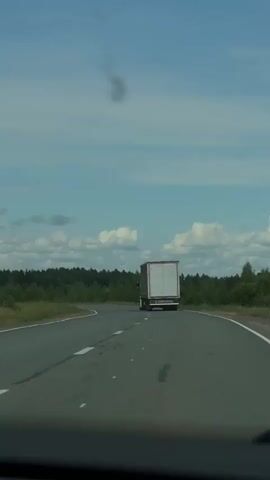 A semi-truck travels down a two-lane road with trees lining the horizon. The sky is blue with scattered clouds. The truck appears to be white with a red stripe on the back. The road has white dashed lines separating the lanes. The camera is positioned inside another vehicle, looking out through the windshield. There is a dark, blurry smudge in the sky above the truck, possibly a bird or debris.