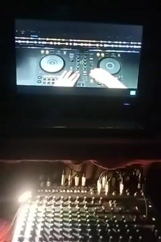 The video shows a close-up, low-angle shot of a DJ mixer in the foreground, illuminated by a bright light. In the background, a laptop screen displays a DJ setup with two turntables and a mixer. A person's hands are visible on the laptop screen, operating the virtual DJ equipment. The overall environment appears to be dimly lit, possibly a club or a performance space.
