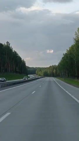 The video shows a view from a moving vehicle on a paved highway. The highway is flanked by dense green forests on both sides, with tall trees dominating the landscape. The sky above is overcast with gray clouds, with a small patch of lighter sky visible in the distance. Two cars are visible on the highway, one in the left lane and another further ahead in the right lane, both moving in the same direction as the camera. The highway has clear white lane markings. The overall scene suggests a journey through a natural, wooded environment.