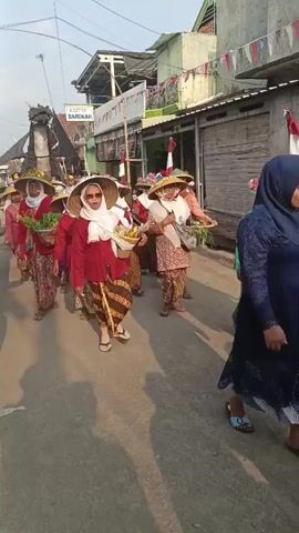 A group of women, dressed in traditional Indonesian attire, are participating in a procession down a street. The women are wearing wide-brimmed hats, red tops, and patterned skirts. Many of them are carrying baskets filled with produce, such as leafy green vegetables and bananas. One woman in the front is wearing sunglasses and a white headscarf. Behind the women, a large, black dragon effigy is being carried. The street is lined with buildings, some of which are decorated with Indonesian flags and colorful banners. The sky is visible overhead, suggesting it is daytime. The overall scene appears to be a cultural celebration or festival.