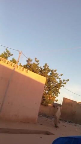 The video shows a street in a neighborhood with buildings, fences, and trash piles. A person wearing a blue hat rides a bicycle past a gate. Then, the camera pans across a building with metal bars in front of it, revealing people inside and a parked motorcycle. The camera continues to pan, showing a large pile of trash and several people in the distance.
