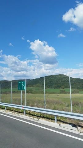 The media file captures a scenic drive on a highway in Capu Piscului, Romania. The scene is set under a bright blue sky dotted with fluffy white clouds, suggesting fair weather and daytime. The road itself is asphalt, marked with white lines, indicating a well-maintained highway. On the right side of the road, a silver metal guardrail runs along the edge, separating the highway from a fence. This fence, made of metal poles and mesh, runs parallel to the road and stretches into the distance, bordering a field. Beyond the field, rolling hills covered in lush green vegetation create a picturesque backdrop. The overall atmosphere is peaceful and expansive, evoking a sense of journey and open road. There is no visible text in the media file.
