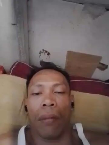 A close-up shot shows a man lying down, facing the camera. He is wearing a white tank top and appears to have earbuds in both ears. The background consists of a rough, textured wall with some visible damage and a cardboard piece leaning against it. There are pillows behind the man's head, one red and one yellow. The lighting is somewhat dim.