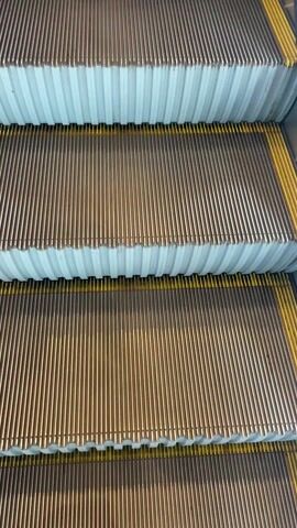 The video starts with a close up shot of an escalator then pulls back to show a person stepping onto it. The camera then moves up the escalator, revealing a large advertisement for a brand featuring three men. The camera continues up the escalator, showcasing the interior of what appears to be an airport, with shops and people visible.