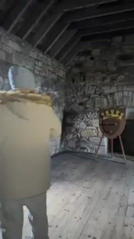 An individual in a tan hooded jacket and gray pants throws a dart at a wooden target in a stone room. The target, designed like a shield, features a crown and already has several darts embedded in it. The room has exposed stone walls and a wooden floor and ceiling, suggesting an old building. The lighting is dim, casting shadows around the space. The person throws three darts, hitting the target with the last two.