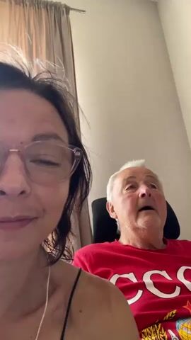 A close-up selfie-style video captures two people. In the foreground, a woman with glasses and dark hair, wearing a thin strap top, smiles faintly with her eyes closed. She appears to be filming. To her right, an older man with short white hair, wearing a red t-shirt with white lettering that appears to say "CCCP" and a graphic featuring a hot air balloon and a globe, looks off to the side with his mouth slightly open. The background shows a light-colored wall and a curtain. The lighting is soft.