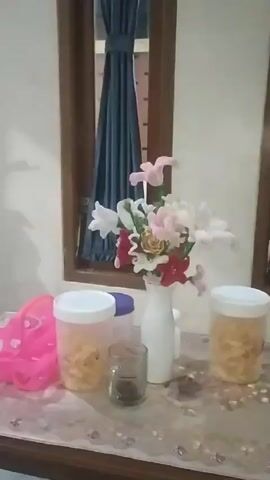 The video shows a still image of a table with several objects on it. In the center of the image is a white vase filled with artificial flowers in various colors including pink, white, and red. To the left of the vase is a pink plastic object, possibly a toy or a decorative item. Next to the pink object and the vase are two clear plastic containers filled with a yellow, crunchy snack. Between these containers and the vase is a small, clear glass with a dark liquid inside. The table is covered with a patterned tablecloth. In the background, a window is visible with a dark blue curtain drawn partially open. The overall setting appears to be indoors, possibly in a home.
