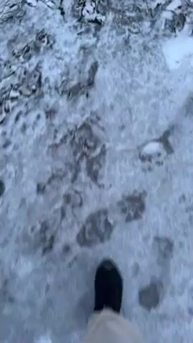 A person wearing light-colored pants and black boots walks through a snowy landscape. The ground is covered in snow, partially obscuring vegetation underneath. The camera is positioned looking downward, capturing the person’s movement from their perspective. The person is stepping over patches of snow-covered brush and foliage.