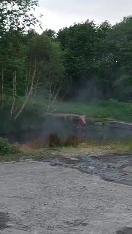 A person is visible standing in a body of water. The person appears to be nude and is facing away from the camera. The water has steam rising from it, indicating it is hot. The surrounding environment is overgrown with trees and grass. A dirt road with puddles of water is visible in the foreground. A dark-colored SUV with a roof rack is parked on the side of the road.