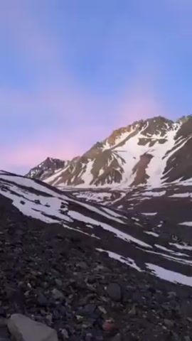 The video shows a mountain range with snow and rock formations under a blue and pink sky. The foreground consists of dark, rocky terrain, and patches of snow are scattered throughout the scene. The mountains in the background are partially covered with snow, and their peaks are jagged. The camera pans slowly to the right.