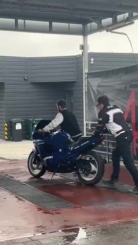 Two men are pushing a blue Suzuki motorcycle across a wet, red surface with drainage grates. The motorcycle is facing left. The man on the motorcycle is wearing a white and black jacket, and the man pushing the motorcycle is wearing a white, black, and gray jacket. The background features a modern gray building with a covered awning. The scene appears to be at a motorcycle wash or service area.