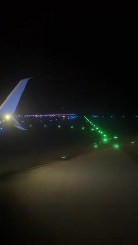 The video captures a night view from an airplane window as it moves along a runway. The wing of the plane is visible on the left side, illuminated by a light. The runway is lined with lights, some blue and some green, indicating its path. In the distance, more lights suggest a city or airport complex. The overall scene is dark, with the runway lights providing the primary illumination. The airplane appears to be either taxiing or preparing for takeoff or landing.