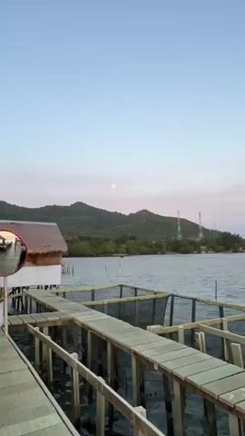 The video opens with a wide shot of a body of water with a pier in the foreground. In the distance, there are hills and a clear sky with the moon visible. The camera then pans to the right, showing a wooden structure with a thatched roof and lights. There are food preparations on a counter. The camera continues to pan, revealing a boat with a thatched roof and seating areas. People are seen disembarking from the boat onto a wooden deck, where many pairs of shoes are scattered. The scene ends with a view of the water and the sky, which is transitioning to sunset colors.