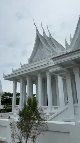 The video shows a white temple building with columns and a traditional Thai roof. The building is painted entirely white, including the walls, columns, roof, and architectural details. The temple is framed by a cloudy sky and stands amidst some greenery. There is a small tree in the foreground near the temple, and a white arched entrance is visible on the left side of the frame. The architectural style includes layered, pointed roofs common in Thai temples, decorated with subtle carvings. The camera appears to be at ground level, looking up at the temple.