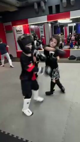 A group of children wearing protective head gear and boxing gloves practice martial arts in a gym. They are punching, kicking and moving around on the mats. One child is wearing white leg protectors. There is a large mirror on one wall. There is a banner for MNT Martine on another wall.