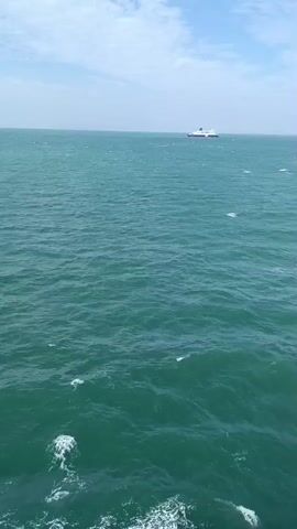 The video shows a wide shot of a body of water. The water is a deep teal color with small waves and whitecaps. In the distance, a white ship with a red stripe is visible. The sky is mostly blue with some scattered white clouds. The camera is positioned as if on another moving vessel, as indicated by the wake visible in the lower portion of the frame. The overall impression is a calm but active sea.