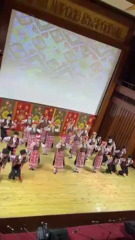 A wide-angle shot depicts a folk dance performance on a wooden stage. A group of approximately 30 dancers, wearing traditional Bulgarian costumes with dark pants, patterned vests, and red-and-white headdresses, are executing synchronized movements. The stage background is decorated with a patterned wall hanging. Above, a large projection screen displays a colorful, abstract pattern. Microphones stand at the edge of the stage. The camera captures the scene from a high angle, looking down at the performers. Sounds of music and cheers are audible.