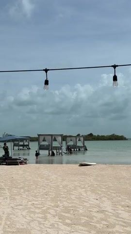 A wide shot of a beach with several cabanas in the water and a string of lights overhead. The sky is partly cloudy and the water is calm. There are a few people in the water near the cabanas. The sand is light-colored and there are a few paddleboards on the beach.