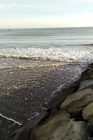 A video of waves rolling onto a dark sand beach. The water is choppy with white foam. In the distance, there is a calm sea with faint horizontal lines of waves and a small boat. The horizon is visible and clear. Rocks are piled along the right side of the frame, forming a breakwater or edge. The sky is overcast and bright.