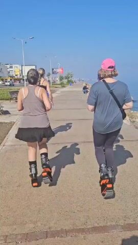 Three people, two women and one person in the distance, are jogging on a paved walkway next to the sea on a sunny day. Two women in the foreground are wearing "Kangoo Jumps" rebound shoes. The woman on the right is wearing a pink cap, a blue T-shirt, and black leggings. The woman in the middle is wearing a grey sleeveless top, a black skirt, and black shorts underneath. The person in the distance is wearing a black sports bra and black shorts. Two people are swimming in the clear blue water. The sky is bright blue and clear.