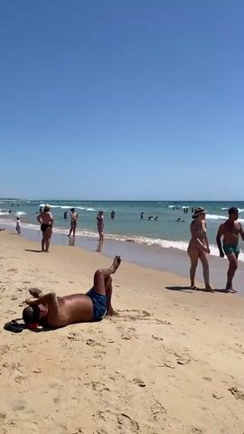 The video opens with a man lying on his back on a sandy beach, facing the ocean. His legs are bent and his arms are relaxed. The camera then pans to the right, showing a couple walking along the shoreline. The woman is wearing a bikini and a hat, while the man is in swim trunks. They are walking towards the water. As the camera continues to pan, more people are visible in the ocean, swimming and playing in the waves. There are several red buoys floating in the water. The sky is clear and blue, indicating a sunny day. The waves are moderate, and the water appears calm. The beach is wide and sandy, with some scattered seaweed. The overall scene depicts a typical day at the beach with people enjoying the sun, sand, and sea.