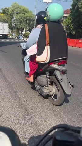 The video shows two people riding on a scooter on a sunny day. The person in front is wearing a blue shirt and red pants, with their bare feet visible. They are wearing a black helmet. The person in the back is wearing a black garment that covers their head and body, and a light blue helmet. A beige bag is slung over their shoulder. The scooter is black and red. They are on a paved road with visible lane markings. In the background, there are trees, buildings, a white truck, and red barriers. The camera appears to be mounted on another vehicle, possibly a motorcycle, looking forward.