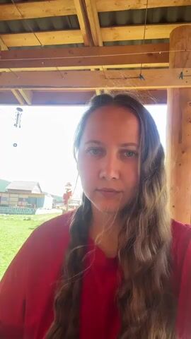 A woman in a red t-shirt with long, wavy, brown hair is looking at the camera, as she adjusts the hair on the right side of her face. She is standing on a porch or under a covered shelter with exposed rafters. In the background, there is a green lawn and several small wooden buildings. A bird feeder hangs from one of the rafters.