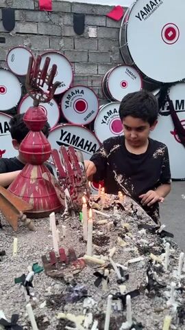Two young boys are the focus of this media file, engaged in an activity involving lit candles. They are positioned in front of a wall adorned with numerous Yamaha drums of varying sizes, some stacked horizontally and others vertically. The drums are predominantly white with red accents and the Yamaha logo. A brick wall forms the backdrop, and some red fabric banners are visible at the top.

In the foreground, on a table or surface covered with what appears to be dirt or ash, are many lit white candles, some partially burned, and some extinguished stubs. Amidst the candles, there are also metal objects that resemble stylized hands and some dark, frayed materials. One boy, wearing glasses and a dark shirt, is partially visible on the left, looking towards the candles. The other boy, in a black t-shirt with a distressed pattern, is actively touching a lit candle with what seems to be a small metal implement. His expression appears focused. The scene is lit by natural light, suggesting it is daytime. The presence of the drums and the candles, along with the somber mood suggested by the candles and dark materials, hints at a ritualistic or commemorative event. The word "YAMAHA" is clearly visible on multiple drums.