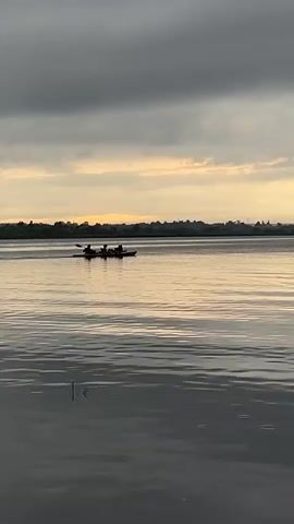 A kayak with three people is visible in the middle distance on a body of water. The sky above is overcast with some areas of lighter clouds and a hint of sunset colors. The water is calm with ripples. The overall scene suggests an outdoor activity, possibly at dusk.