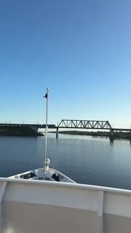 A ship moves forward on a wide river under a clear blue sky. The bow of the ship is visible in the foreground, with a flagpole extending upward. In the distance, a large truss bridge spans the river. A train with several cars is crossing the bridge. The riverbanks are lined with trees. The water is calm.