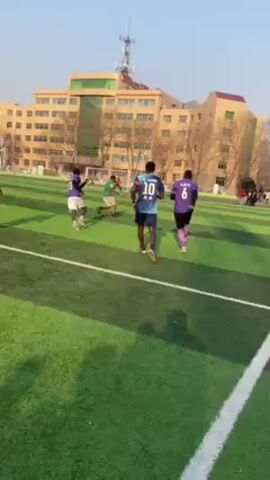 Several people are running on a green turf field. They are wearing athletic clothing. There are buildings visible in the background. The camera is moving and focused on the ground.