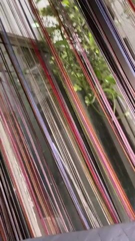 The video shows a close-up, angled view of a string curtain. The strings are thin and appear to be made of a shiny material, with a variety of colors including white, gray, black, red, pink, orange, and yellow. They hang vertically in front of a window, partially obscuring the view of lush green foliage outside. The sunlight filters through the strings, creating a shimmering effect. The camera is handheld and moves slightly, causing the strings to sway gently.
