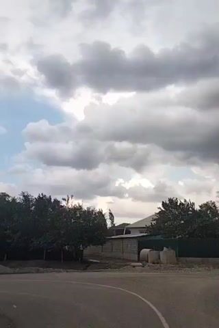 The video shows an outdoor scene on a cloudy day. The camera pans across a street and a row of trees. The trees are dense and green, and their branches are swaying in the wind, indicating a breezy or stormy atmosphere. In the background, there's a house with a white roof and a fence made of concrete blocks. Some construction materials, possibly pipes or tanks, are visible near the fence. The sky is filled with large, gray clouds, suggesting impending rain or a transition in weather.
