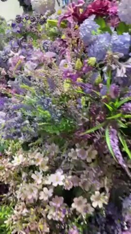 The camera pans across a display of artificial flowers. The majority of the flowers are shades of purple, from lavender to deep violet. There are also some white and pink flowers interspersed. The display is dense and creates the impression of a flower wall. The background is out of focus, but it appears to be a store or event space. There is background music playing.