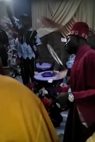 The video appears to be taken from a low angle, looking upwards into a crowded space. Several people are visible, including individuals wearing a red hat and shirt, a black shirt with white writing, and a bright yellow shirt. There are various objects scattered on the floor, including what looks like a decorative white hat with a purple band, fabric remnants, and some floral arrangements. The environment seems somewhat enclosed with striped fabric hanging in the background. Music is playing, and the overall impression is that the people are gathered in this space.