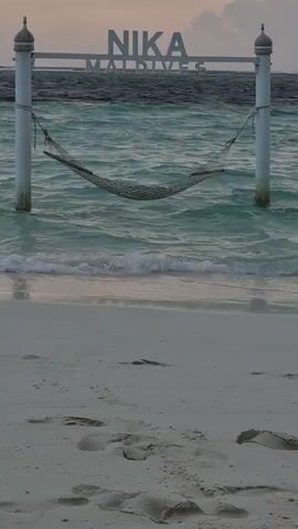 The video depicts a serene beach scene at Nika Maldives. A hammock is strung between two white posts set partially in the water. Waves gently lap at the sandy shore. The Nika Maldives sign is positioned above the hammock. The camera pans right, revealing the ocean and the distant island. The sun is partially obscured by clouds, casting a soft light across the water.