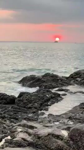 The video shows a rocky coastline at sunset. The sun is a bright red orb on the horizon, partially obscured by a distant island. Waves are gently crashing against the dark, jagged rocks in the foreground. The sky is a soft mix of gray and pink hues. The water is a light grey color, reflecting the sky's color. There are no people visible in the frame.