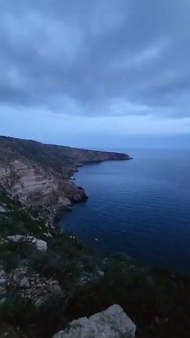 The camera pans across a landscape of a dark blue ocean and rocky coastline on a cloudy day. The perspective is from a high vantage point, overlooking the water and the edge of a peninsula jutting out into the sea. Vegetation grows along the cliffside in the foreground. The sky is overcast with varying shades of gray and blue clouds. A small boat is visible in the distance on the water.