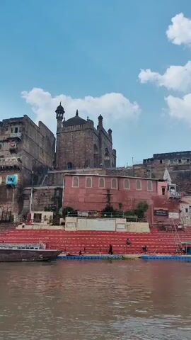 A slow pan across a body of water reveals a large, multi-tiered embankment with red steps leading up to several buildings. A boat is partially visible on the left side of the frame. The buildings are constructed of brick and stone, with some appearing older and more weathered than others. One prominent building in the center features a dome and two minarets, suggesting it is a mosque. Another building, painted pink, has a row of arched windows. Figures can be seen on the steps and at the base of the buildings. The sky is blue with scattered white clouds. The water is murky brown.