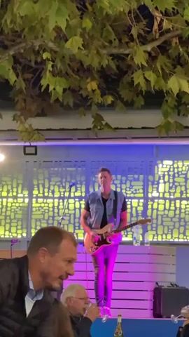 The video shows a man playing an electric guitar on a stage outdoors at night. The man is wearing a denim vest, a black t-shirt, and dark jeans. He is standing in front of a backdrop with a pattern of rectangular cutouts that is lit with yellow light. The stage itself is lit with purple light. In the foreground, there are people sitting at tables, watching the performance. A tree with green leaves overhangs the scene, partially obscuring the view of the stage. A microphone stands in front of the guitarist. There is a suitcase or amplifier on the right side of the stage.
