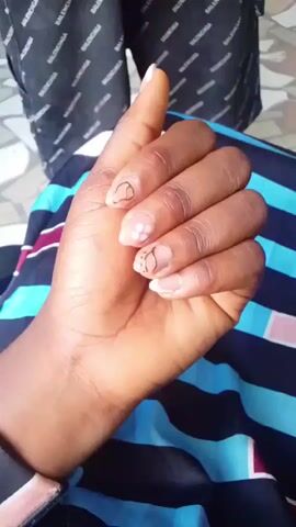 A close-up shot of a hand with acrylic nails, some with gold detailing, is shown. The hand rotates to display all angles of the nails. In the background, a person wearing a Balenciaga shirt and black pants can be seen, along with a tiled floor.