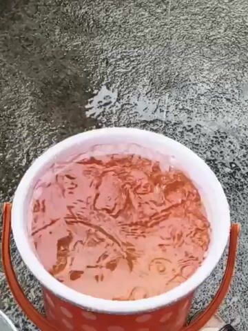 A close-up, overhead shot shows an orange and white polka dot bucket filled with reddish-brown liquid. Raindrops are creating ripples and splashes on the surface of the liquid. The bucket has orange handles. The background is a wet, grey, textured surface, likely pavement.