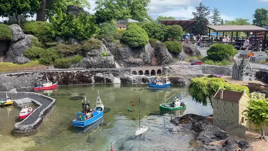 This is a wide shot of a miniature Lego harbor scene. In the foreground, there is a body of water with several Lego boats of various sizes and colors, including a white sailboat, a green fishing boat, and a blue fishing boat. There are also buoys in the water. To the left, a curved Lego breakwater encloses a small area with two white Lego boats. To the right, there are small Lego buildings resembling stone houses and a castle. In the middle ground, there is a rocky hillside with Lego trees and bushes. A Lego train track runs along the base of the hill, with a small red Lego train visible on the track. There are Lego bridge arches over the water. In the background, there is a covered seating area with people sitting and talking, and a few trees. The sky is partly cloudy. The scene appears to be set outdoors in daylight.