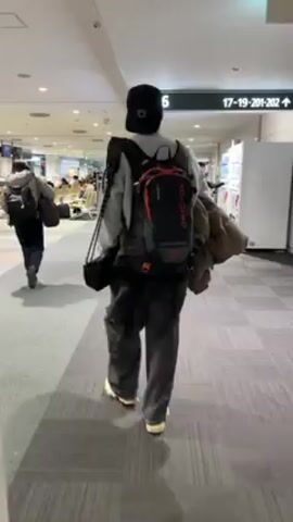 A person with a backpack and a baseball cap is walking through an airport. They are wearing dark pants, a light-colored sweatshirt, and white sneakers. They are carrying a brown coat over one shoulder. There are other people walking in the background, and some are sitting in chairs. There are vending machines on the right side of the screen, and a sign pointing to gate 16 is visible. The floor is gray carpet. The lighting is bright and even.