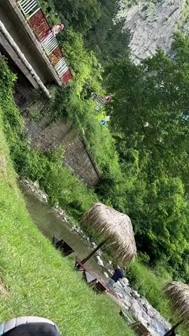 A person wearing a pink hat is standing on a bridge structure. Below the bridge, a shallow stream flows past some rocks. Two thatched umbrellas are set up on the grassy bank near the stream, with lounge chairs visible beneath them. Another person is seated under one of the umbrellas, facing the stream. The background is filled with lush green trees and a steep, rocky incline.